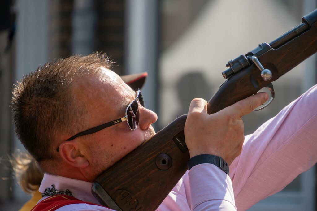 A man with sunglasses aiming a rifle outdoors, focused on precision.